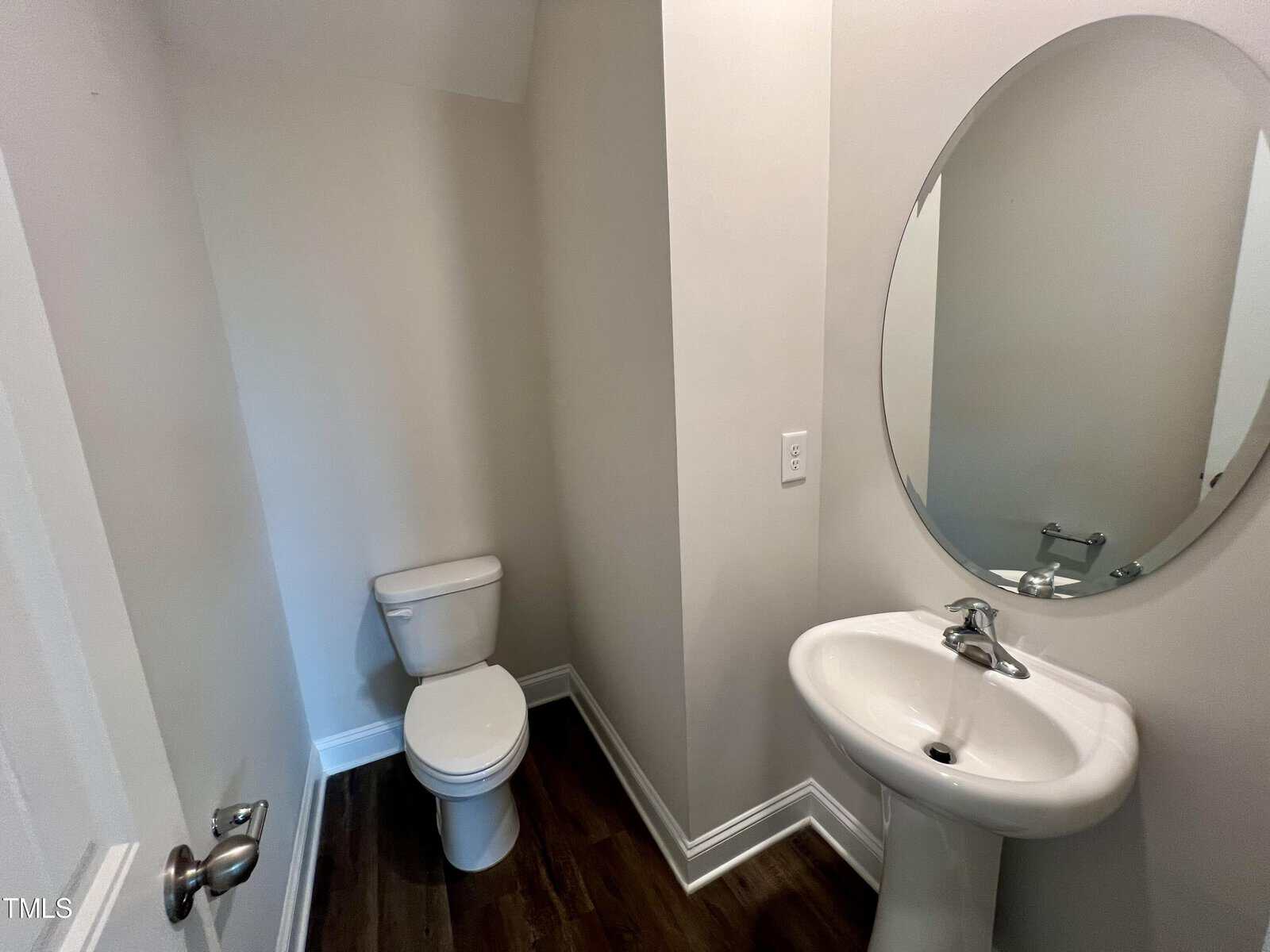 A small bathroom featuring a white toilet and a pedestal sink. An oval mirror is mounted above the sink. The room has white walls and dark wood flooring. A closed door is partially visible on the left side.