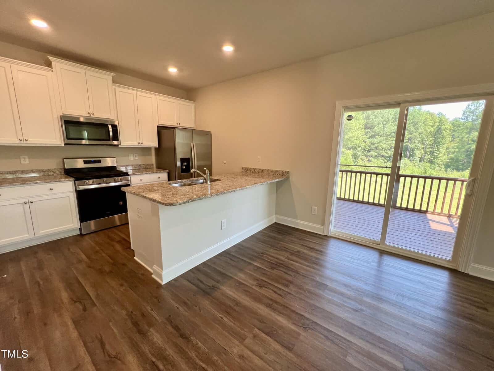 A kitchen with wooden floors, white cabinets, and granite countertops. Includes a stainless steel fridge, oven, and microwave. A sliding glass door leads to a deck with a view of greenery outside. Ceiling lights illuminate the space.
