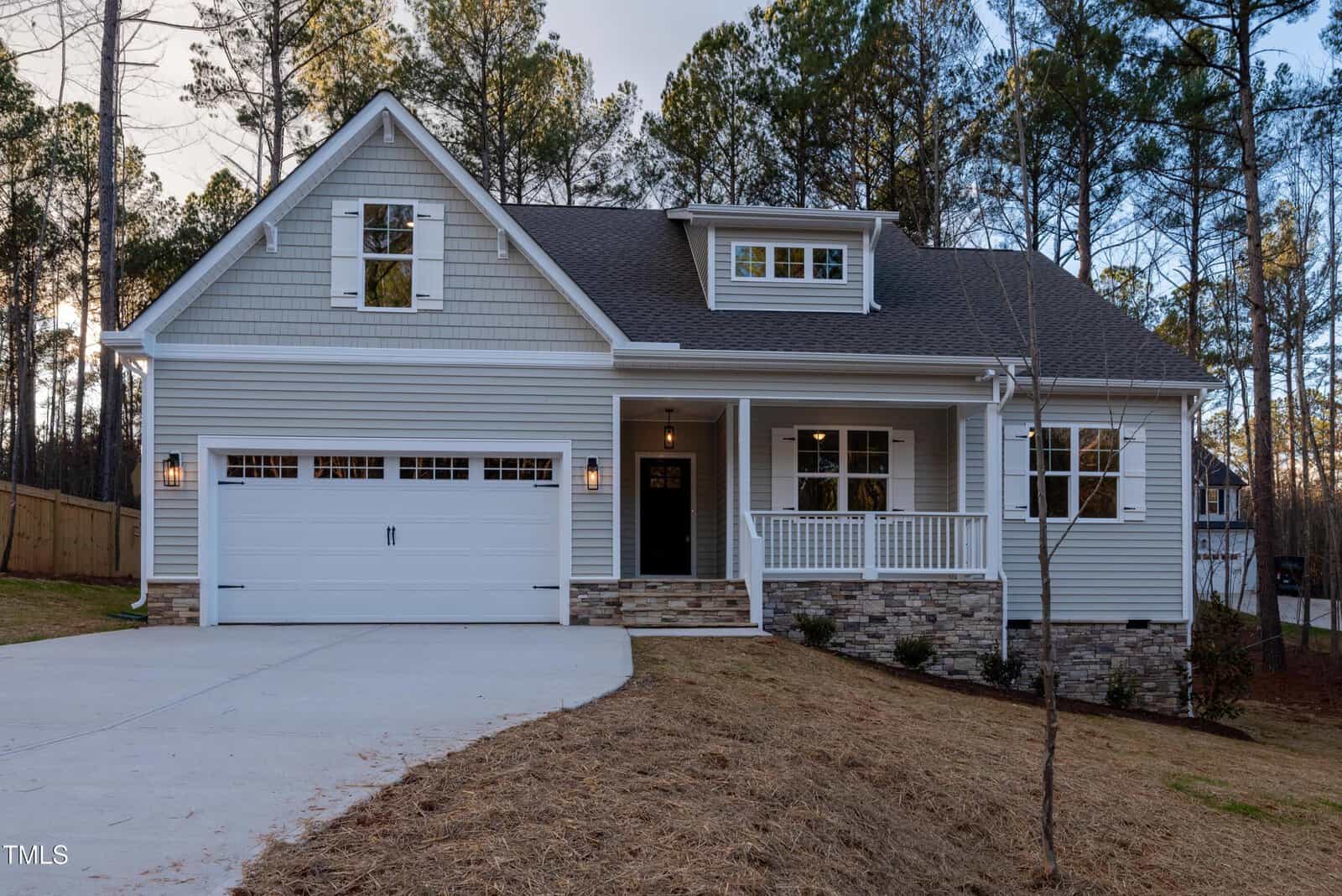 A modern single-story house with gray siding, stone accents, a covered front porch, white trim, and a two-car garage, set against a backdrop of tall pine trees and a sloped driveway.