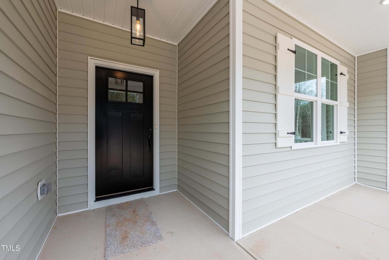 A covered front porch with light gray siding, a black front door with glass panels, a doormat, a lantern-style light fixture, and a window with white shutters on the right.