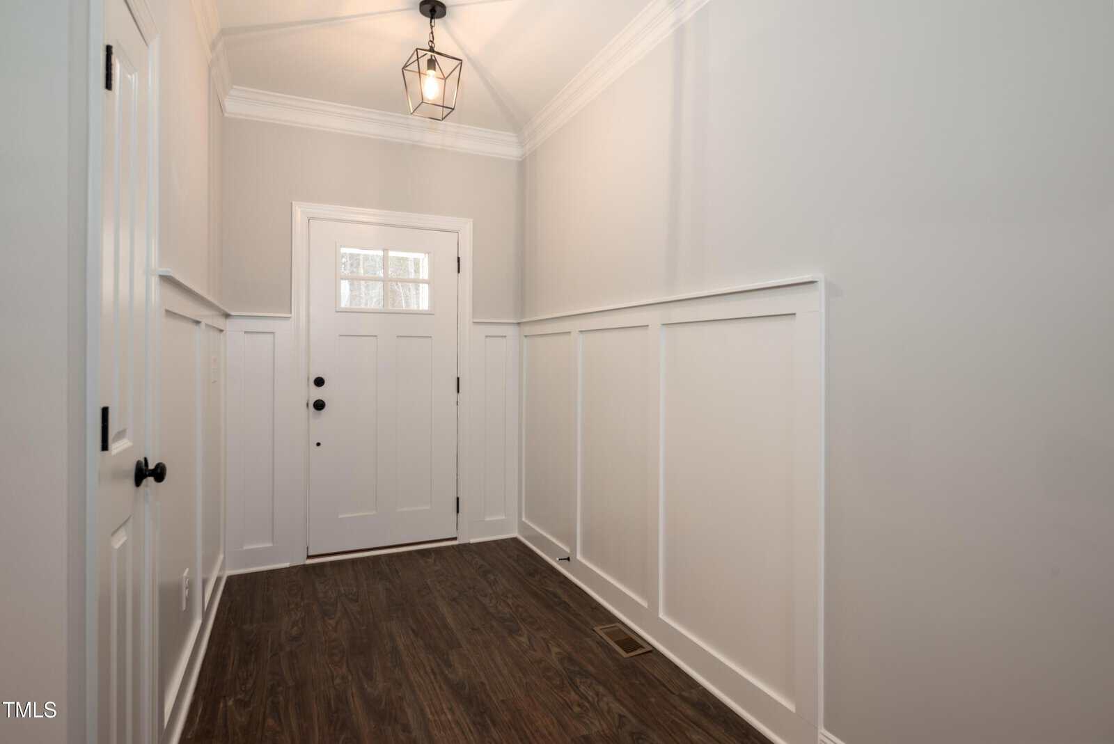 A bright entryway with white walls, wainscoting, a white front door with glass panes, dark wood flooring, and a black geometric pendant light hanging from the ceiling.