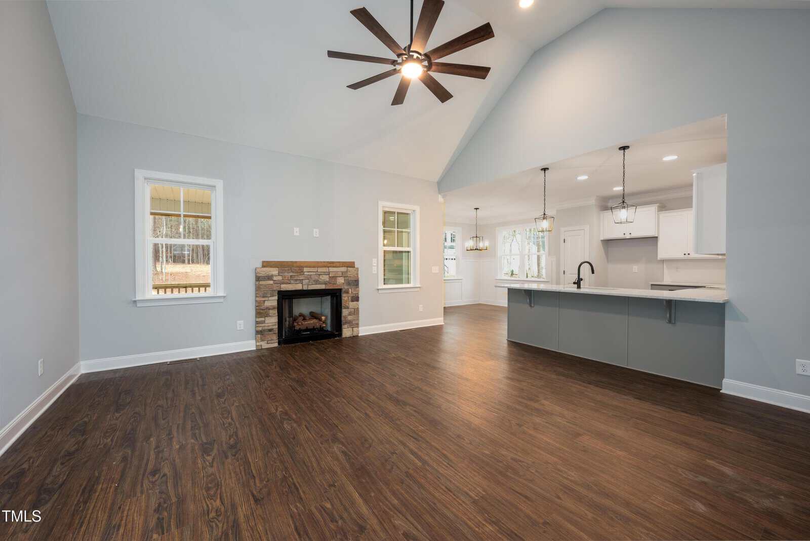 Spacious open-concept living area with wood floors, stone fireplace, ceiling fan, and recessed lighting. The kitchen features white cabinets, pendant lights, and a large island with a sink, all under a vaulted ceiling.