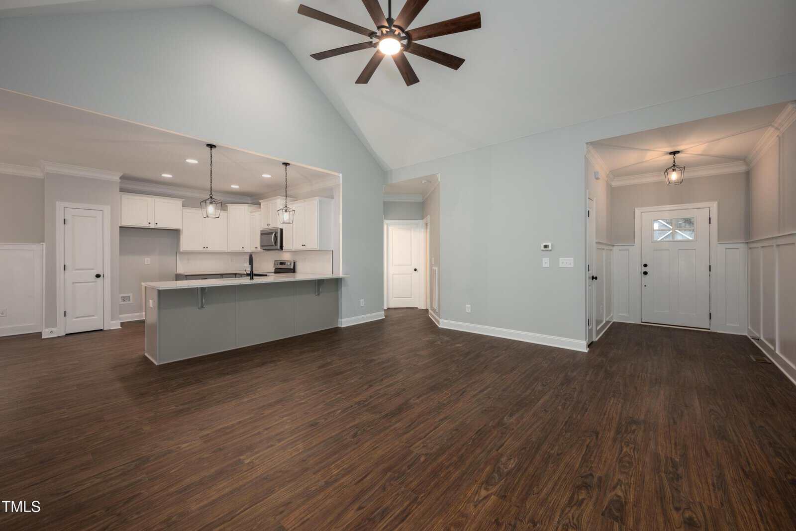 Spacious open-concept living area with vaulted ceiling, dark wood floors, light gray walls, a modern kitchen with white cabinets and stainless steel appliances, and a foyer with a glass-paneled front door.