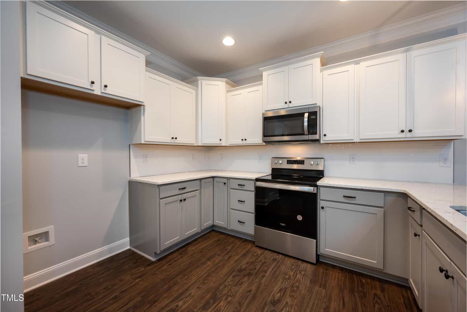 Modern kitchen with white upper cabinets, gray lower cabinets, stainless steel oven and microwave, white countertops, white tile backsplash, and dark wood floors. There’s an empty space likely for a refrigerator.