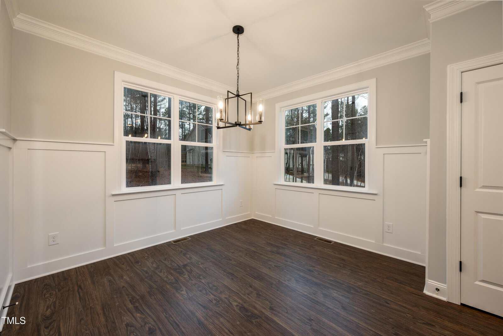 Bright dining room with white paneled walls, dark wood floor, large windows showing trees, and a black modern chandelier hanging from the ceiling.