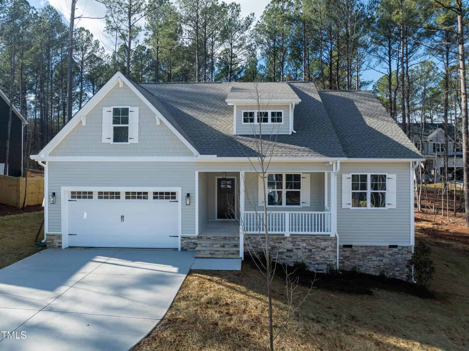 A modern two-story house with light gray siding, white trim, a covered front porch, and an attached two-car garage, set among tall pine trees and a sloping grassy yard.