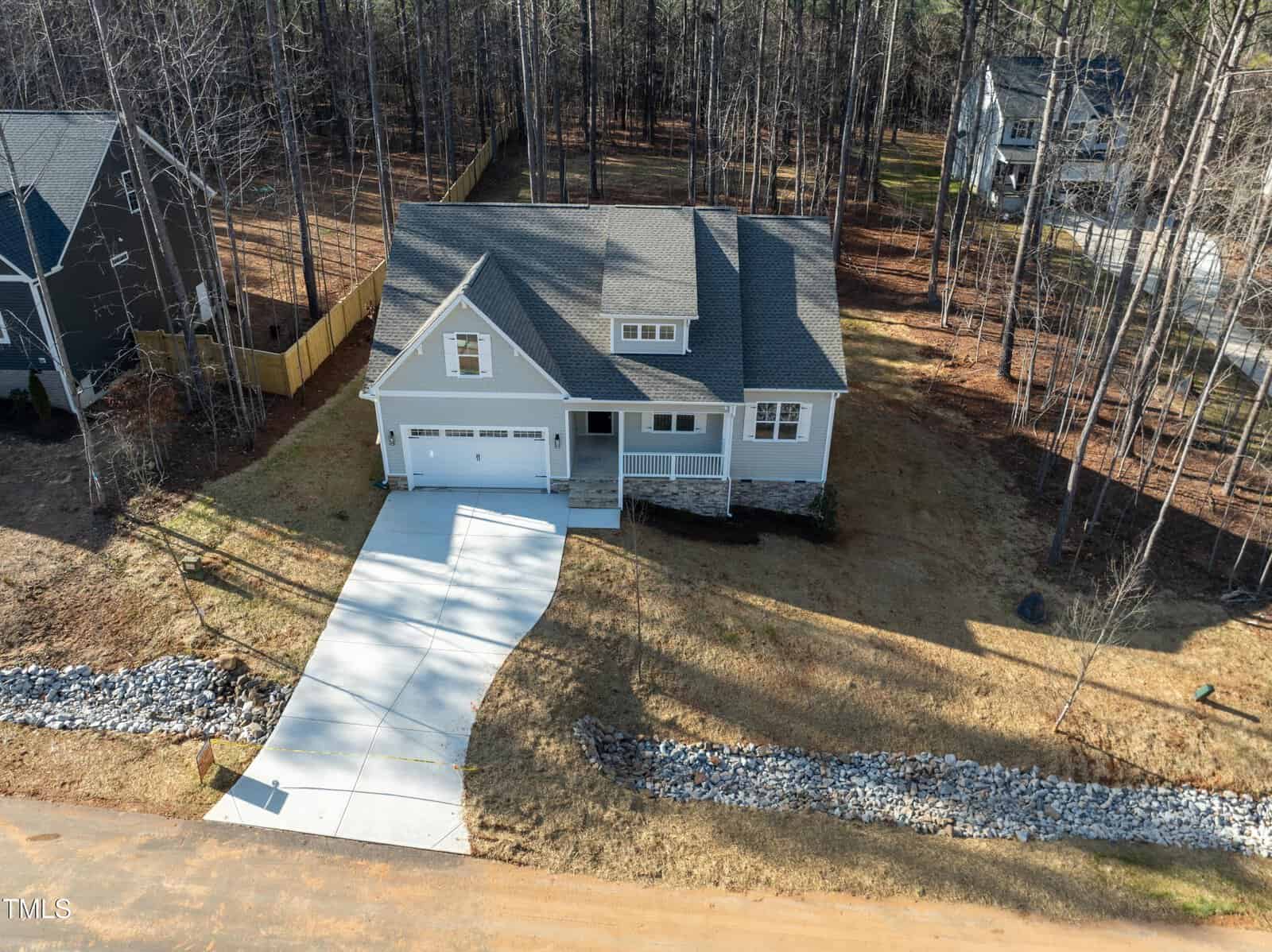 Aerial view of a two-story gray house with a two-car garage, concrete driveway, and lawn. The house is surrounded by tall trees and neighboring homes, with a rocky drainage area beside the driveway.
