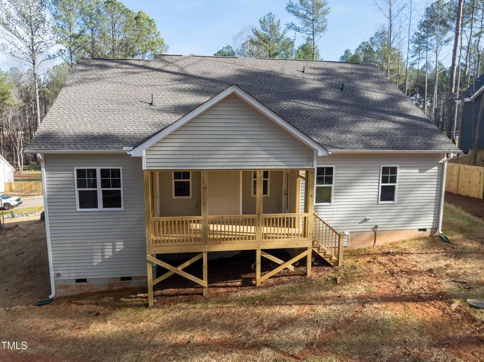 A single-story house with light gray siding and a gabled roof features a covered wooden back porch with railings and steps, set on a dirt backyard surrounded by trees and neighboring homes.