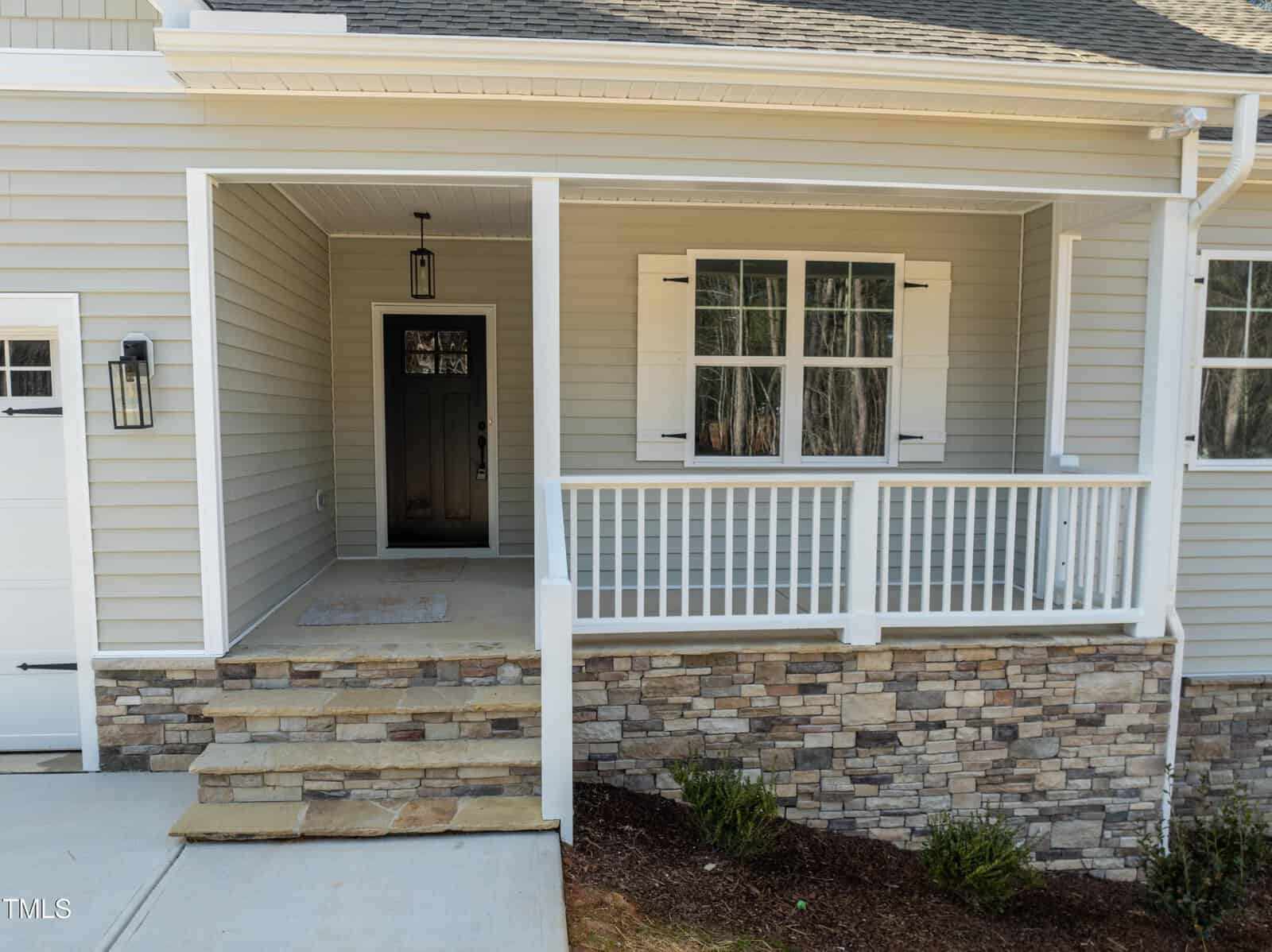 Front porch of a house with beige siding, white railing, stone accents, and a black front door. Steps made of stone lead up to the porch, which has a hanging light fixture above the door. Shrubs are planted along the edge.