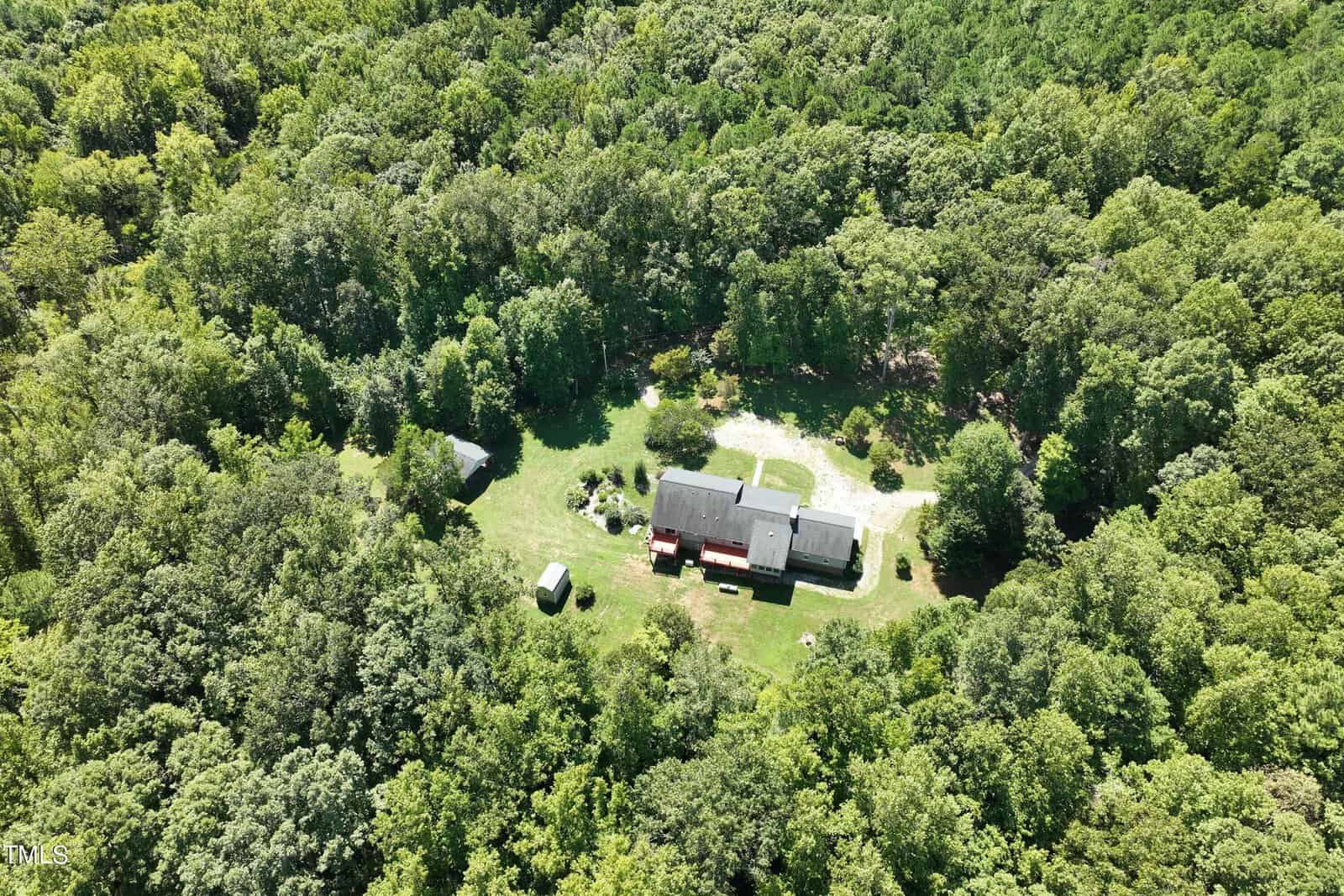 Aerial view of a house at 4161 Fernwood Trail, nestled in lush green forest near Oxford. The main building and two smaller structures are partially shaded by trees, with a grassy clearing and dirt path visible in the foreground.