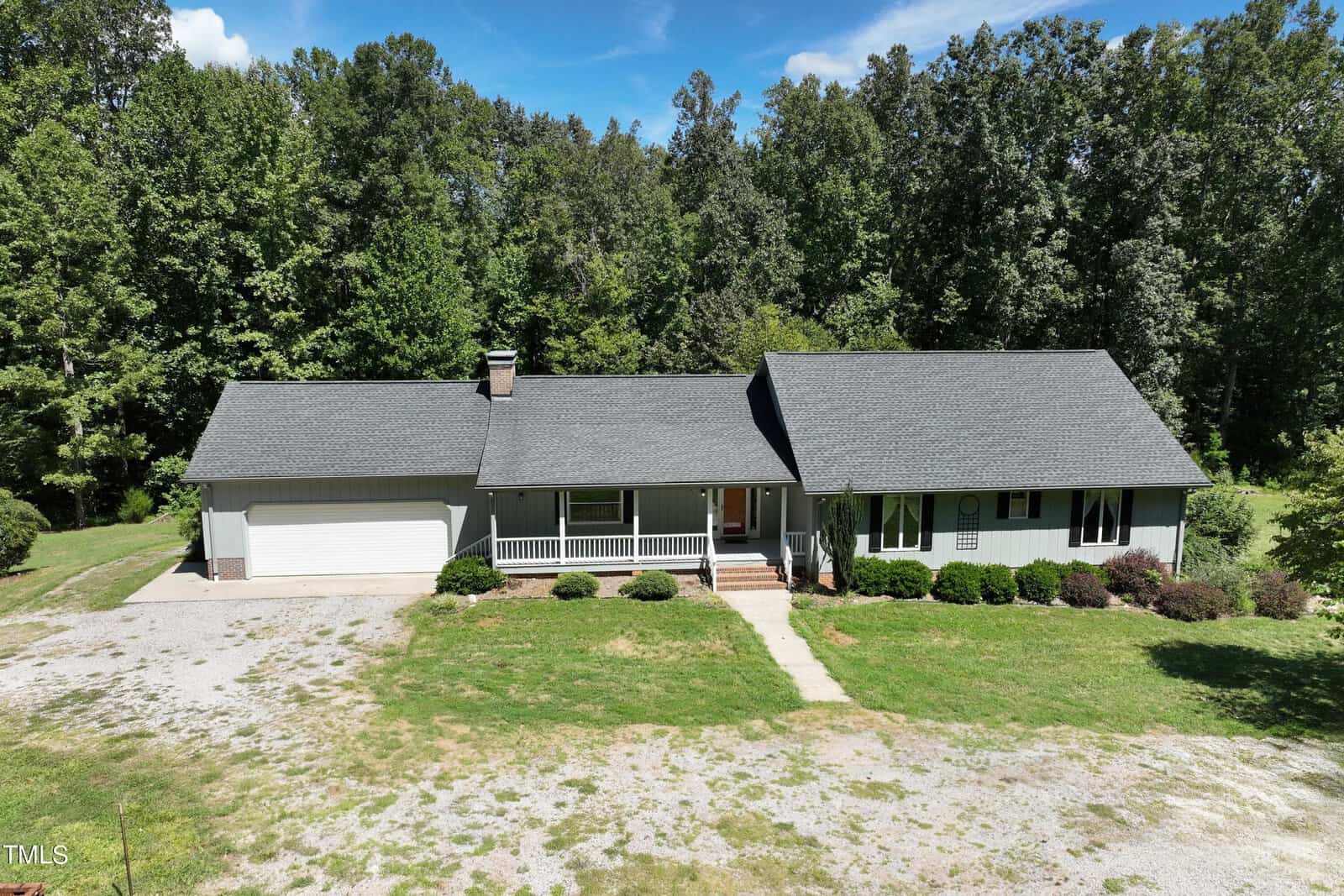 A single-story house with a gray roof and light-colored exterior sits surrounded by lush trees. A grassy lawn and gravel driveway lead up to the front porch. The background features a clear blue sky with some clouds.