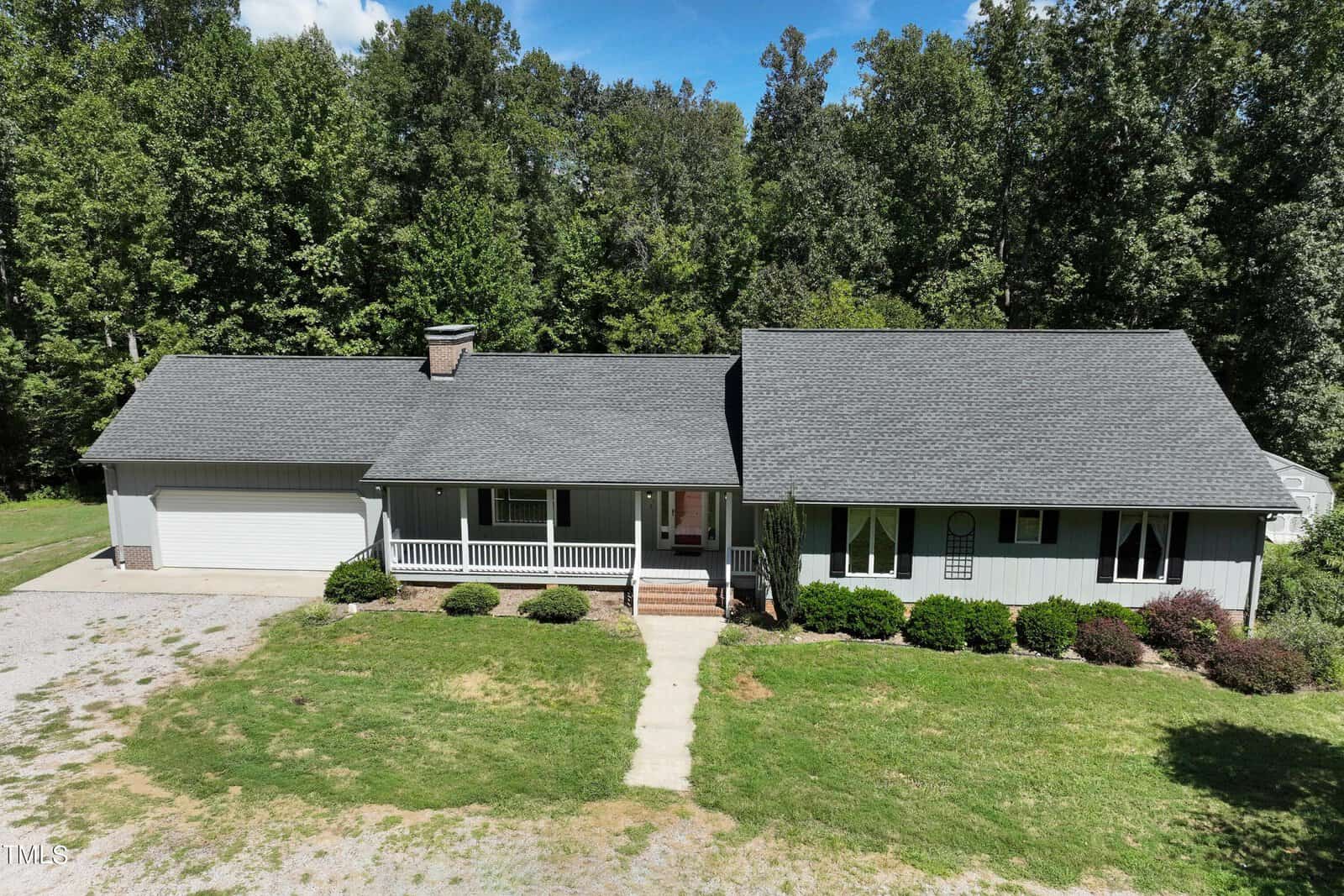 A single-story gray house with a dark roof, surrounded by lush green trees. It has a front porch with white railings and a brick chimney. A concrete walkway leads to the entrance.