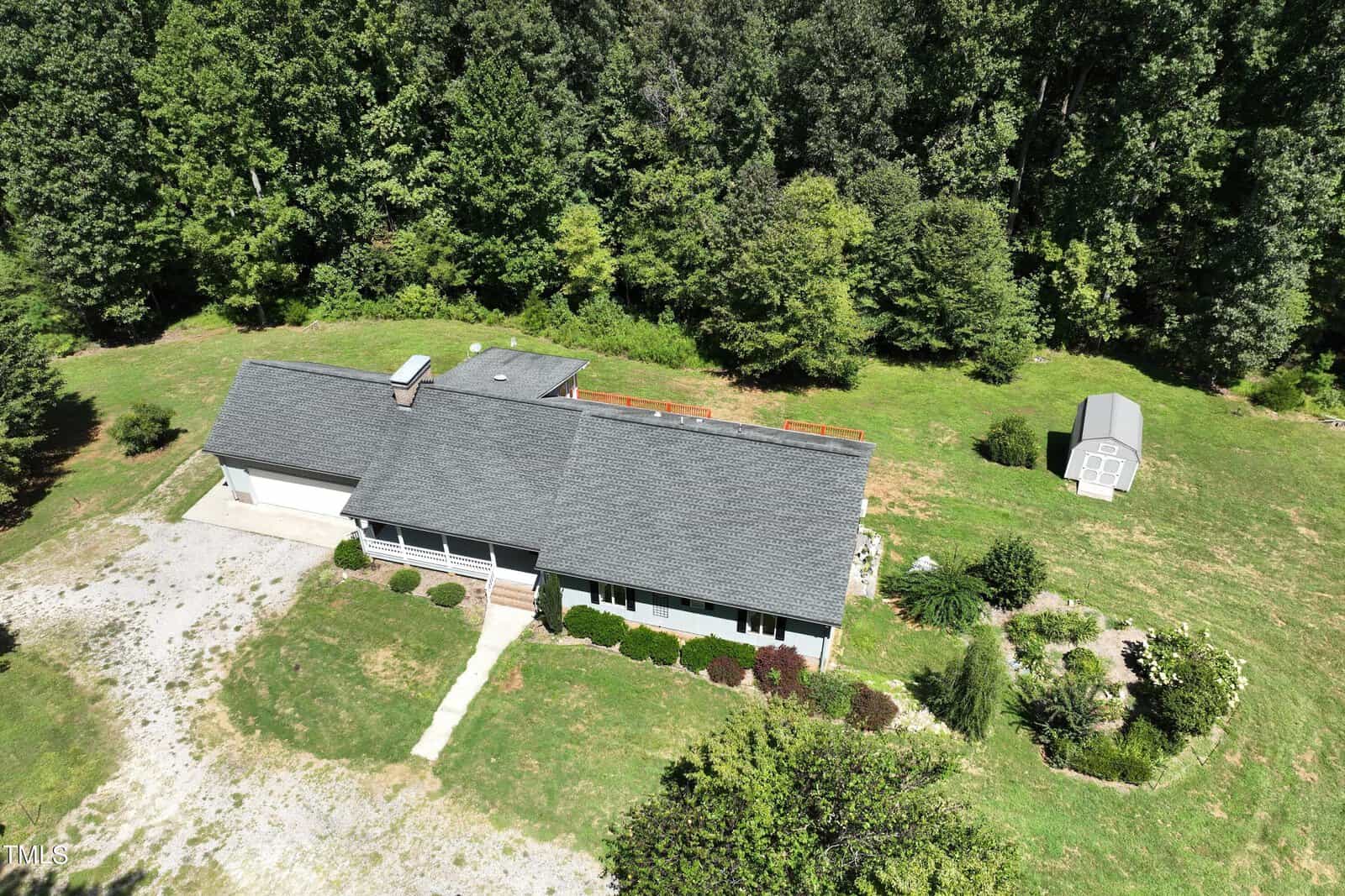 Aerial view of a single-story house with a gray roof surrounded by lush greenery and trees. A small shed is visible in the backyard, and a pathway leads to the front door. The yard is spacious, with a mix of grass and bushes.
