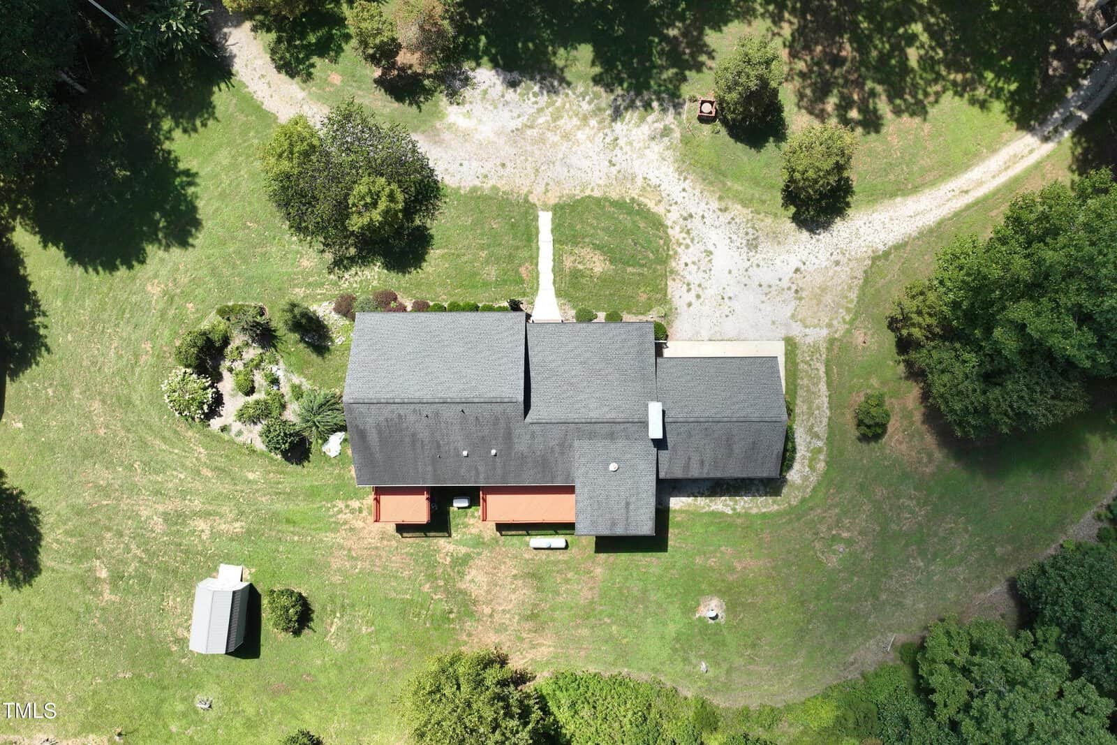 Aerial view of a house with a dark roof, surrounded by green grass and trees. The property includes a small outbuilding and a gravel driveway curving around the front. A pathway leads to the front entrance.