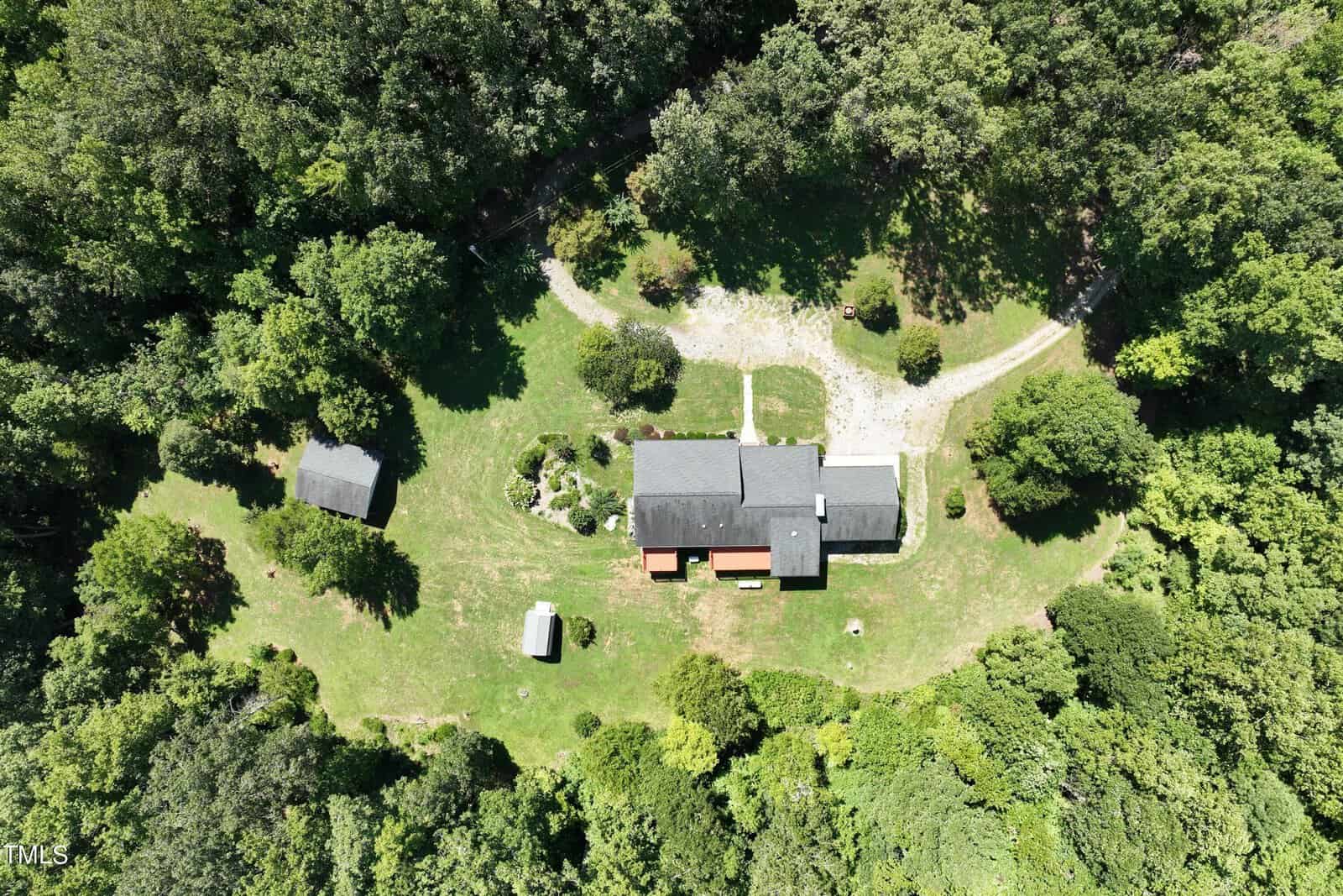 Aerial view of a red and gray house surrounded by a lush green forest. The property includes a small shed and a large yard with a winding gravel path leading to the house. Dense trees encircle the open space.