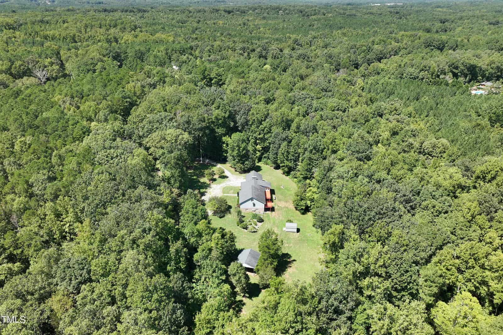 Aerial view of a secluded house surrounded by dense green forest. The property includes a main house, a smaller structure, and a few open spaces in a clearing, with a driveway leading through the foliage.