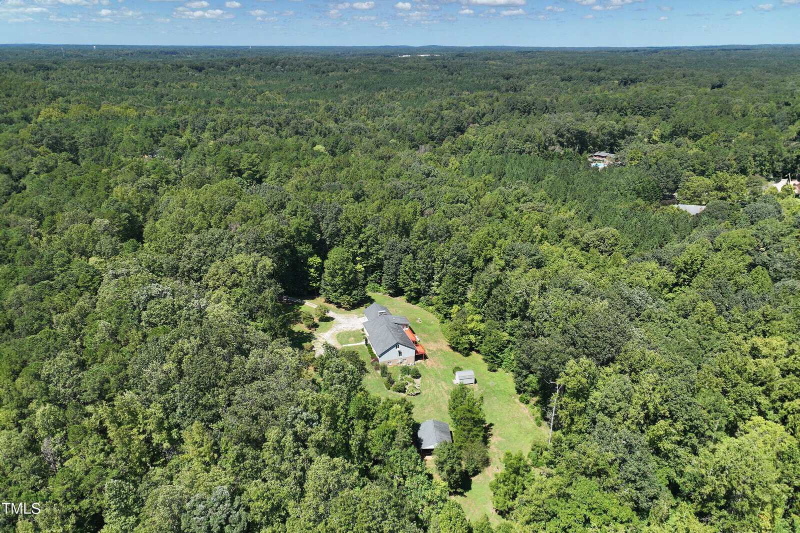 Aerial view of a dense forest with a small clearing featuring a house and two smaller buildings. The landscape is lush with greenery under a blue sky dotted with clouds.