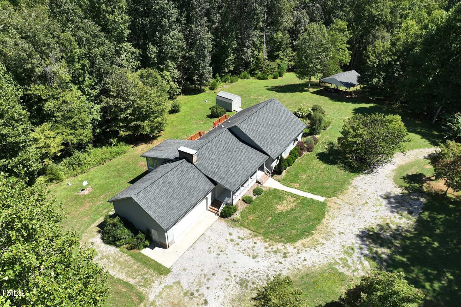Aerial view of a single-story house with a dark roof, surrounded by lush greenery and trees. There is a gravel driveway leading to the garage. A small structure and a gazebo are in the backyard.