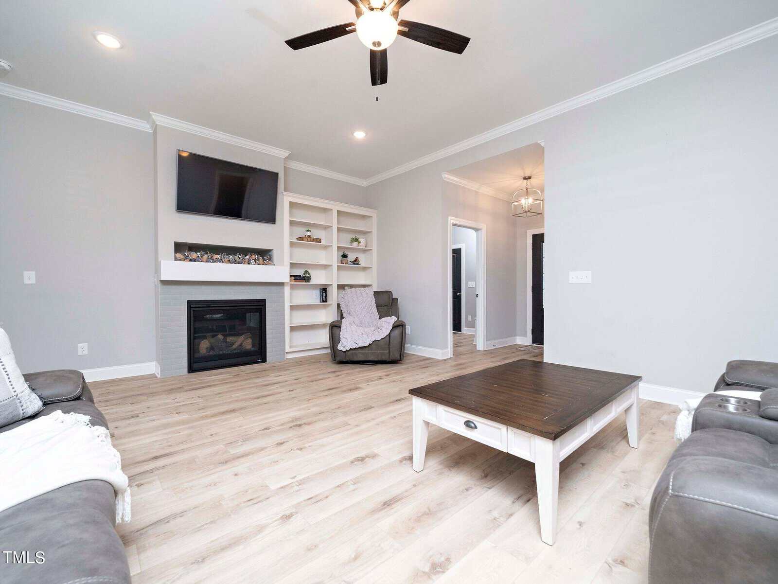 A modern living room with light wood floors, gray walls, and a ceiling fan. It features a fireplace with a wall-mounted TV, built-in shelves, leather armchairs, and a large coffee table in the center.