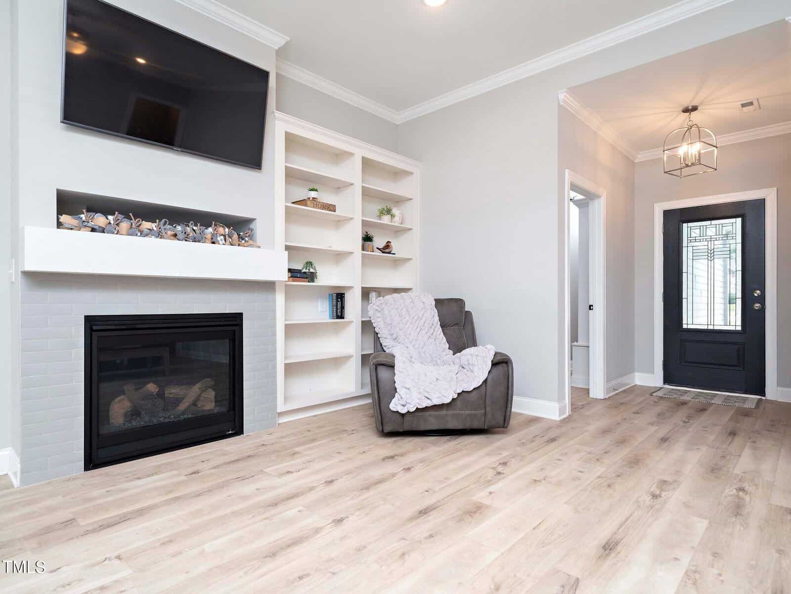 A cozy living room with a fireplace and built-in shelves, featuring a large TV above. A plush armchair with a gray blanket sits nearby. The room has light wood flooring and a dark front door with glass panels, leading to a bright hallway.