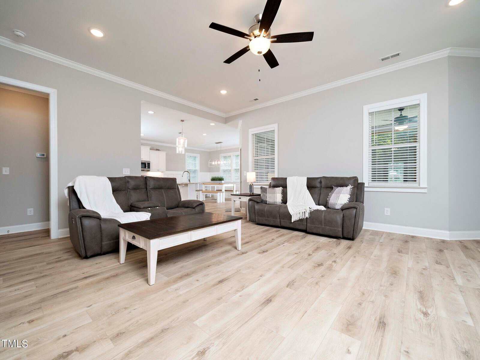 Spacious living room with light wood flooring, two gray sofas with white throws, and a wooden coffee table. A ceiling fan hangs above. The room opens to a bright kitchen with white cabinetry and a central island in the background.