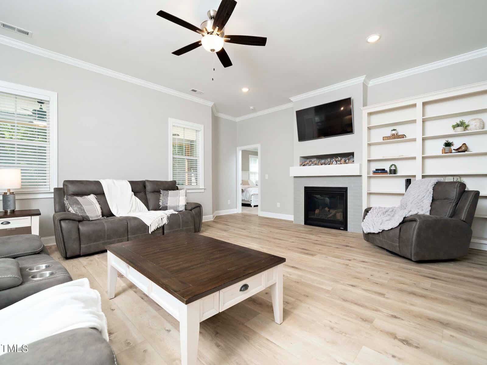 A modern living room with gray sofas and a recliner. A wooden coffee table sits in the center. There is a fireplace with a TV above it, and open shelves on the side. The room has light wood flooring and a ceiling fan.