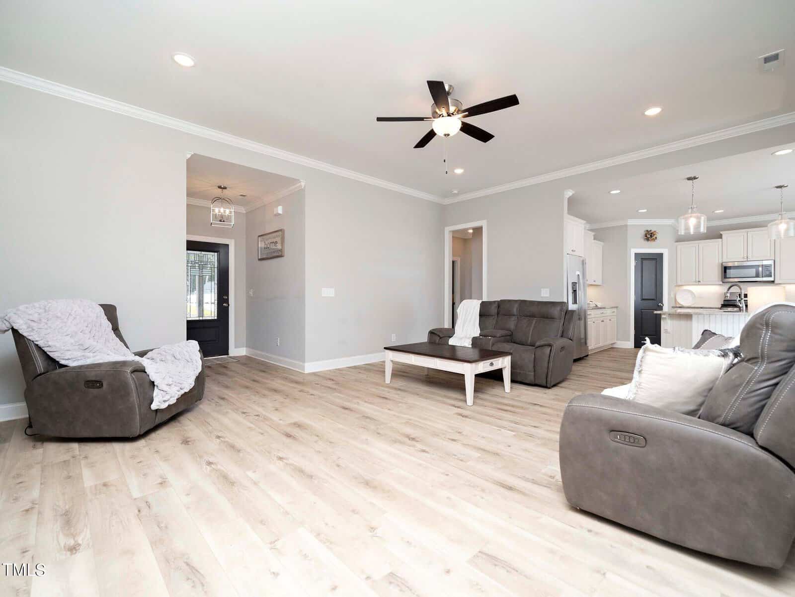 Spacious living room with light wood flooring, featuring two gray recliners, a matching sofa, and a white coffee table. Ceiling fan with lights overhead and an open view into a modern kitchen with white cabinetry and pendant lighting.