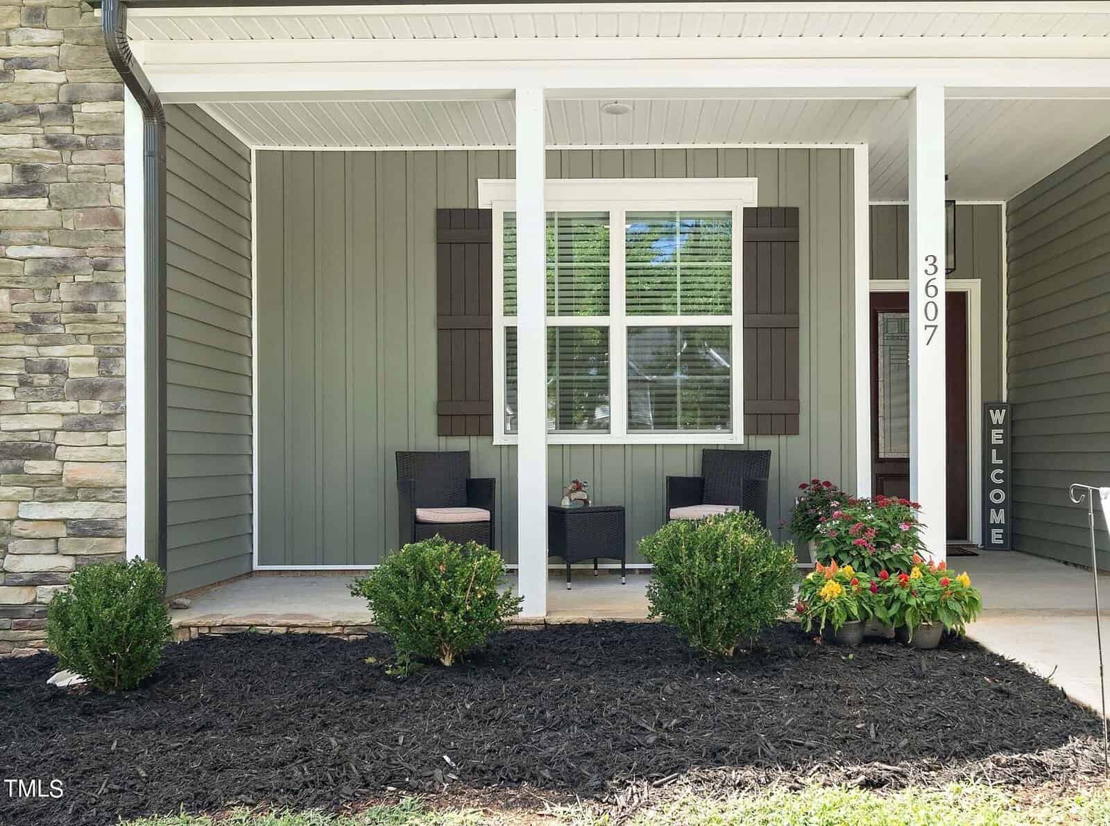A cozy front porch with two woven chairs and a small table. The house has green siding, white columns, and a vertical "Welcome" sign. Potted flowers and shrubs decorate the area, and the number "3607" is displayed on a column.