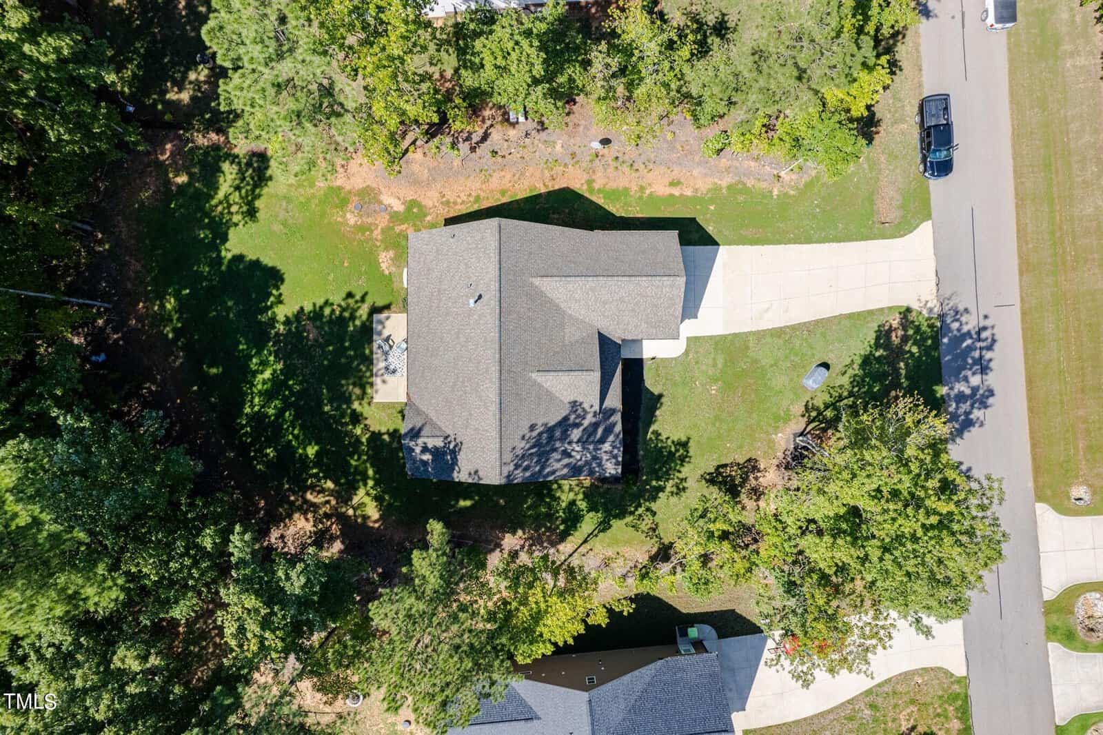 Aerial view of a suburban house with a dark roof, surrounded by trees and a green lawn. The property includes a driveway leading to a garage, and a street with a parked car is visible on the right.