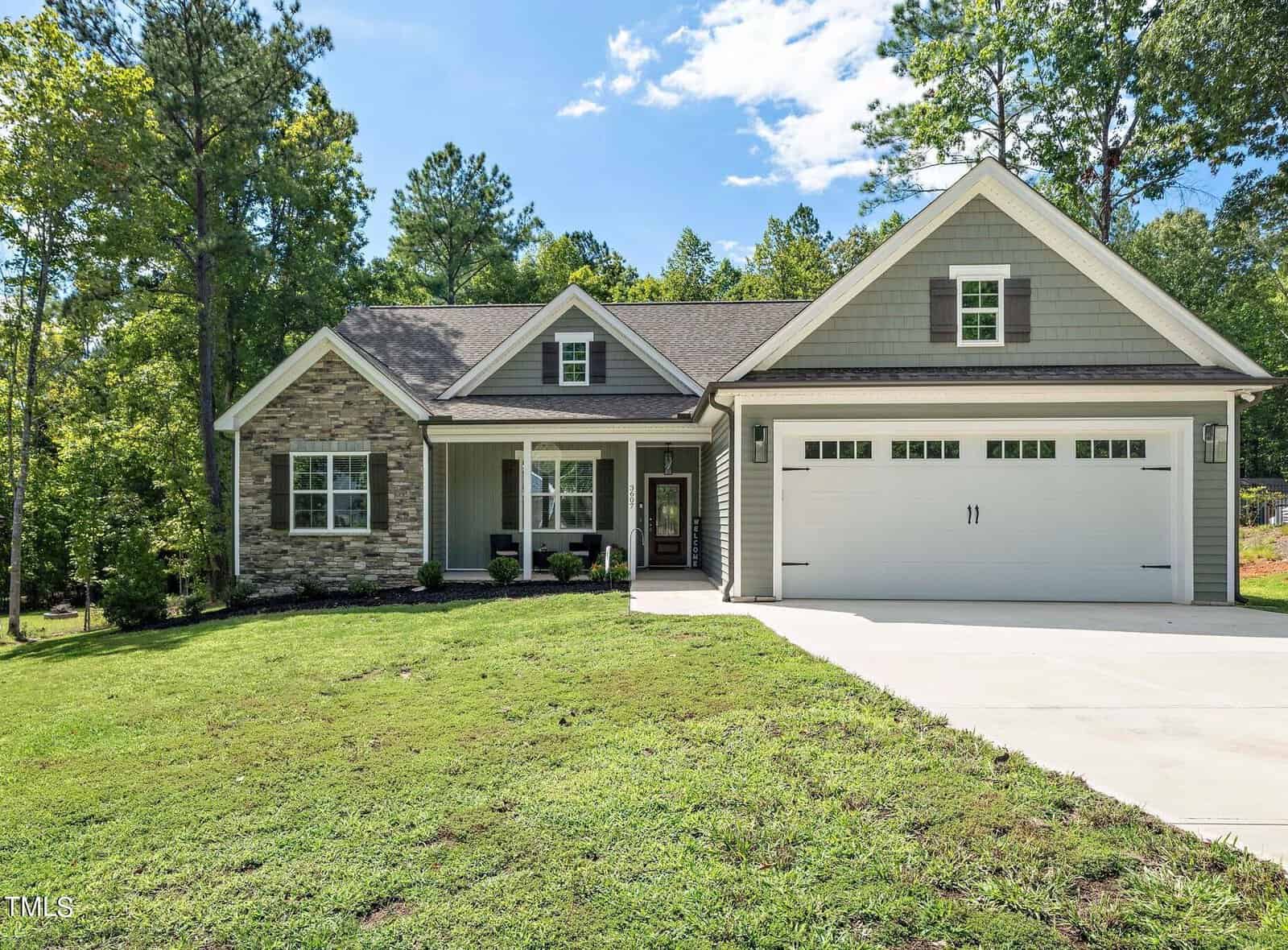 Nestled on 3607 River Watch Lane in Franklinton, this single-story house with gray siding and stone accents is embraced by trees under a partly cloudy sky. It features a covered porch with white columns and a two-car garage, complemented by lush green grass and a paved driveway in the front yard.