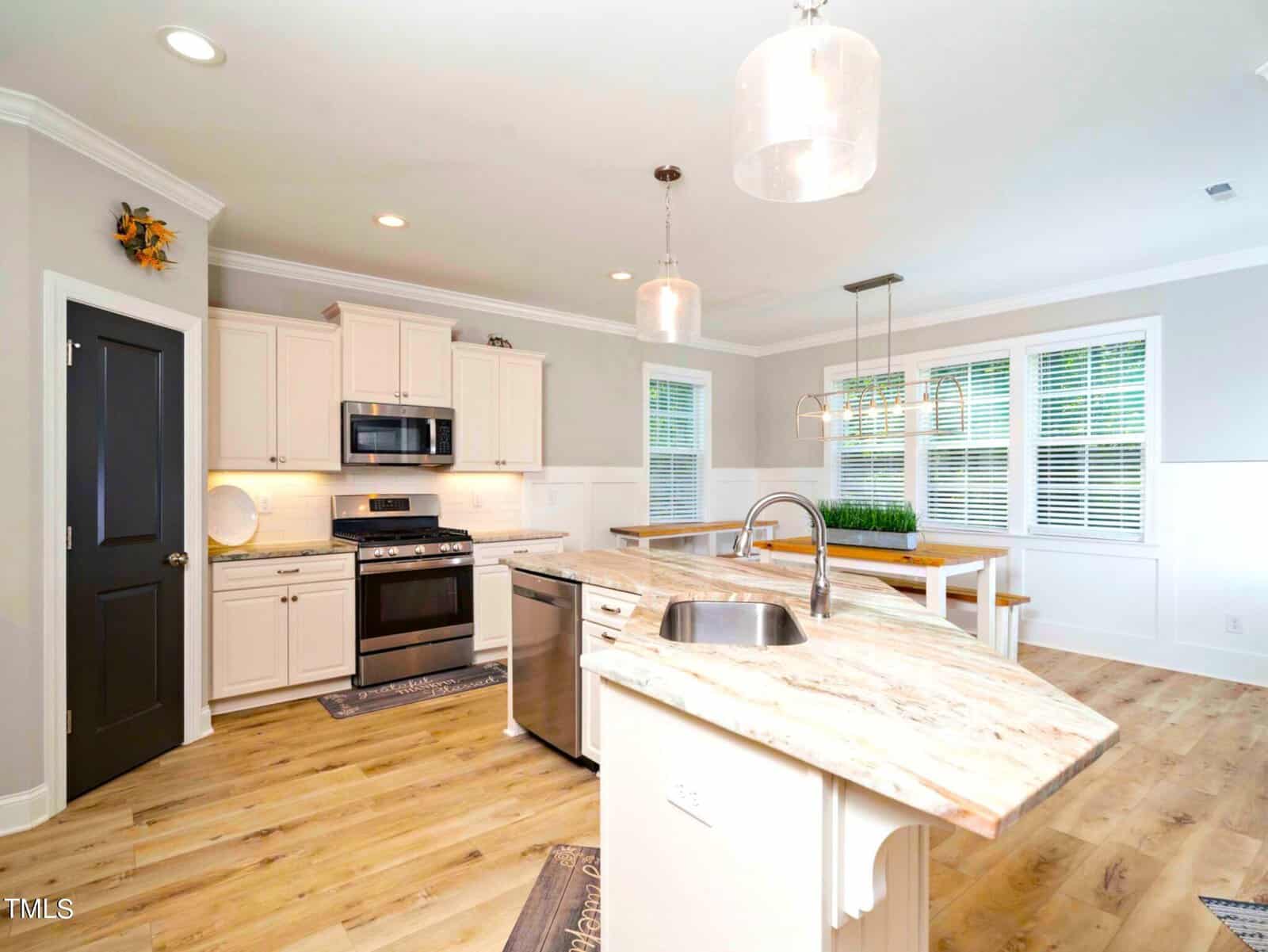 A modern kitchen with white cabinets, a stainless steel oven, and a dishwasher. The kitchen island has a marble countertop and sink. A dining area is visible, featuring a bench and chairs under hanging lights. The floor is light wood.