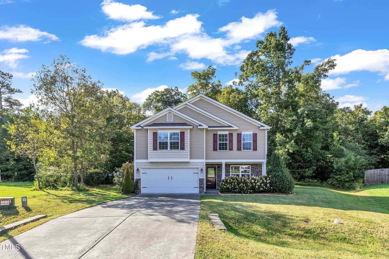 Nestled among trees, this charming two-story house at 1842 Fillmore Drive in Creedmoor features light gray siding and white trim. It boasts a two-car garage and a neatly maintained lawn under a clear sky with a few clouds.