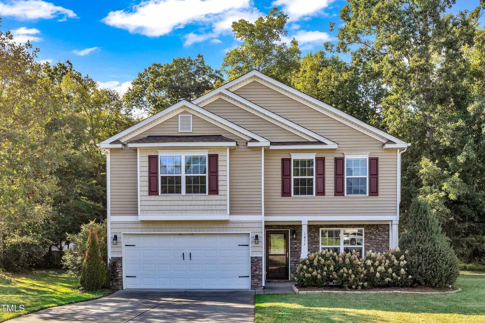 A two-story house with beige siding and maroon shutters stands under a blue sky. It features a white garage door, symmetrical windows, and an entryway with shrubs on either side. Trees surround the property, providing a leafy backdrop.