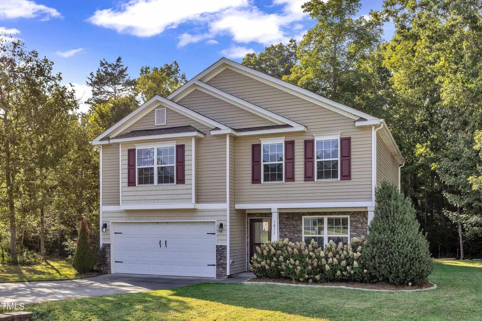 A two-story beige house with brown shutters and a two-car garage is surrounded by well-maintained greenery. It is set against a backdrop of lush trees and a clear blue sky. The driveway and lawn are neatly kept.
