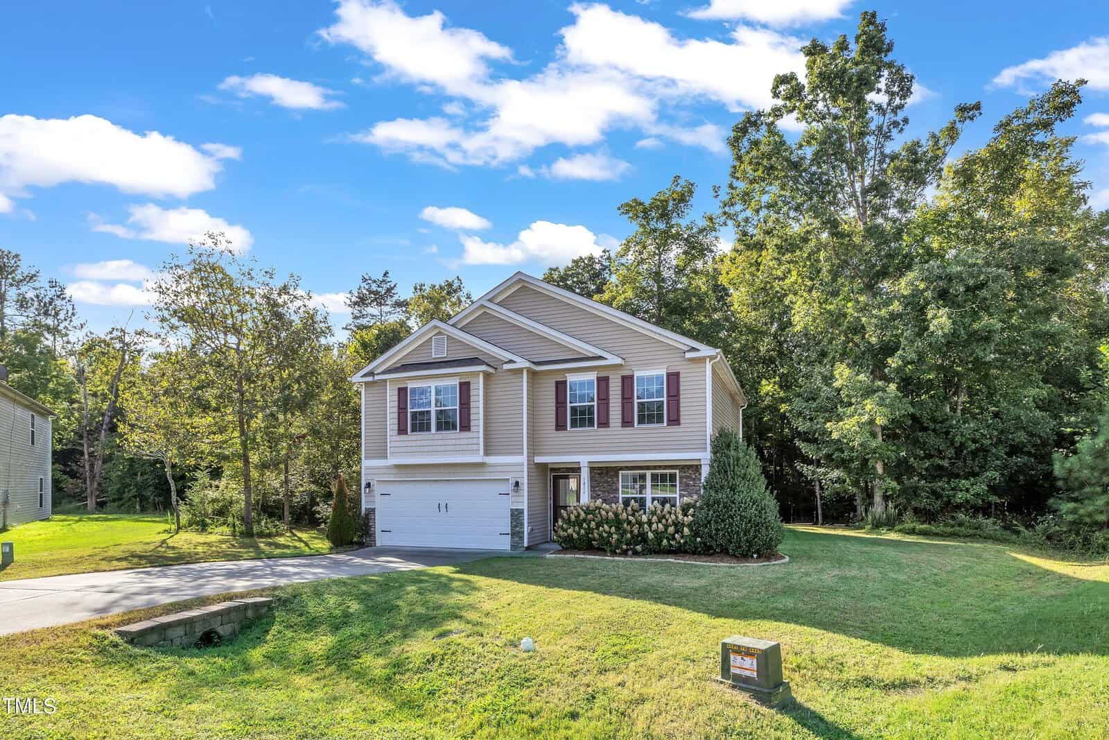A two-story house with beige siding and red shutters, set against a backdrop of trees. A well-maintained lawn and driveway are in the foreground, with a "For Sale" sign near the curb under a clear blue sky.
