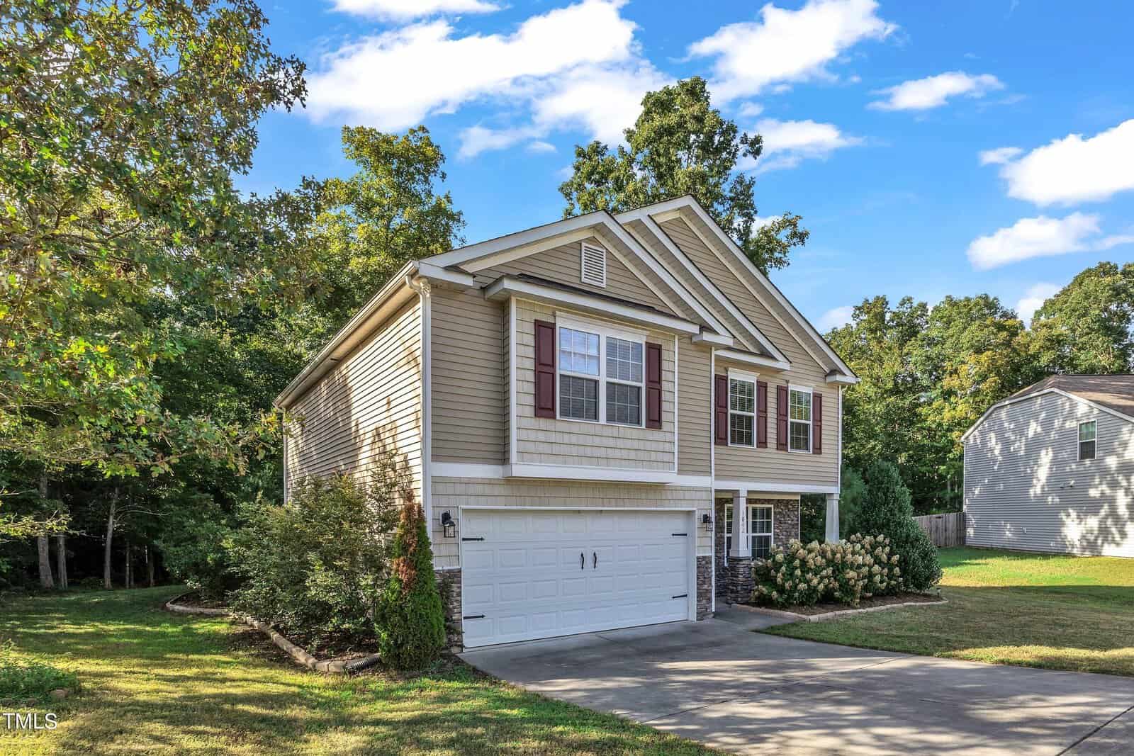 Two-story beige house with white trim and red shutters, featuring a two-car garage. Surrounded by trees and a well-maintained lawn, the house sits under a clear blue sky with scattered clouds.