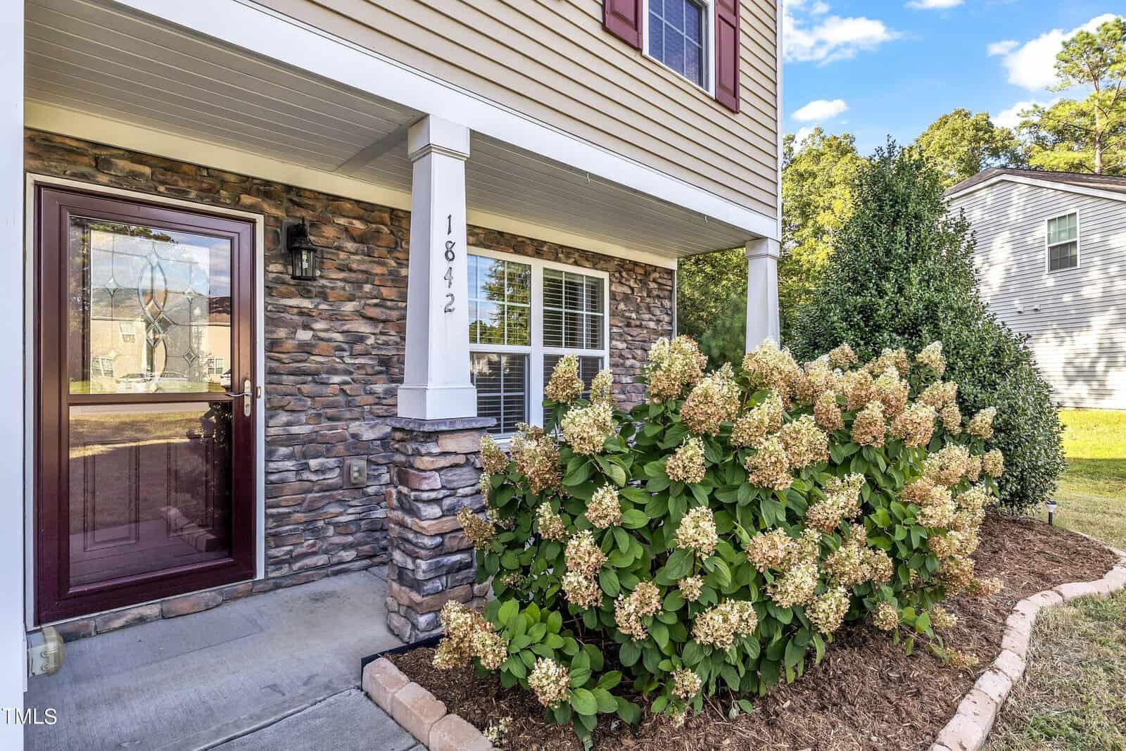 Front view of a house with beige siding and stone accents. A glass front door is beside a large window. Lush hydrangea bushes with white flowers line the walkway, and the house number "1842" is displayed on the wall. Trees are visible in the background.
