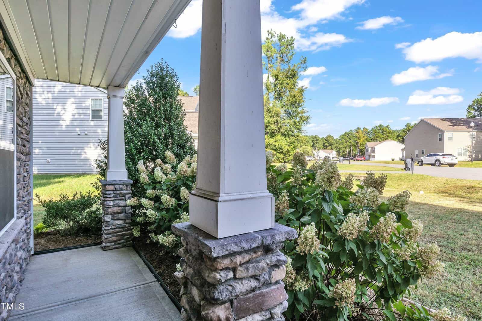 View from a porch with stone pillars, overlooking a garden with lush greenery and blooming hydrangeas. In the background, suburban houses line a well-maintained neighborhood street under a blue sky with scattered clouds.