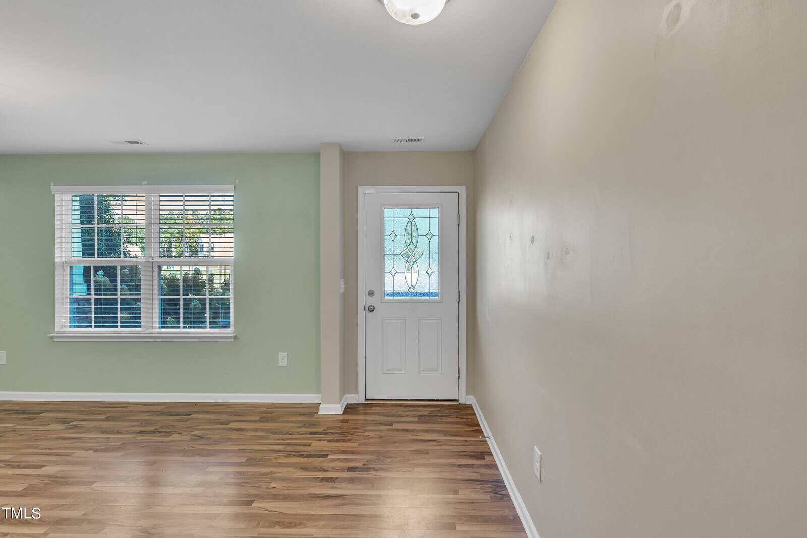 An empty room with light green and beige walls features a window with blinds on the left and a front door with decorative glass on the right. The floor is wood, and a ceiling light fixture is visible.