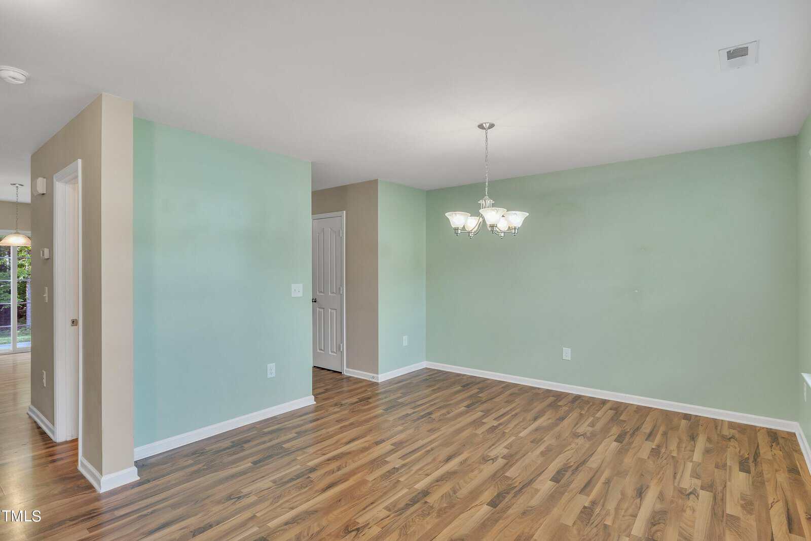 An empty room with light green walls and wood flooring. A chandelier hangs from the ceiling. An open doorway leads to another area, and a closed white door is visible. Natural light comes through a window on the left.