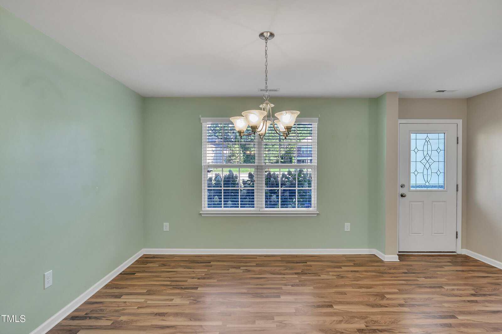 A room with green walls and wooden flooring features a decorative chandelier hanging from the ceiling. A window with blinds allows light to enter, and a white door is seen on the right.