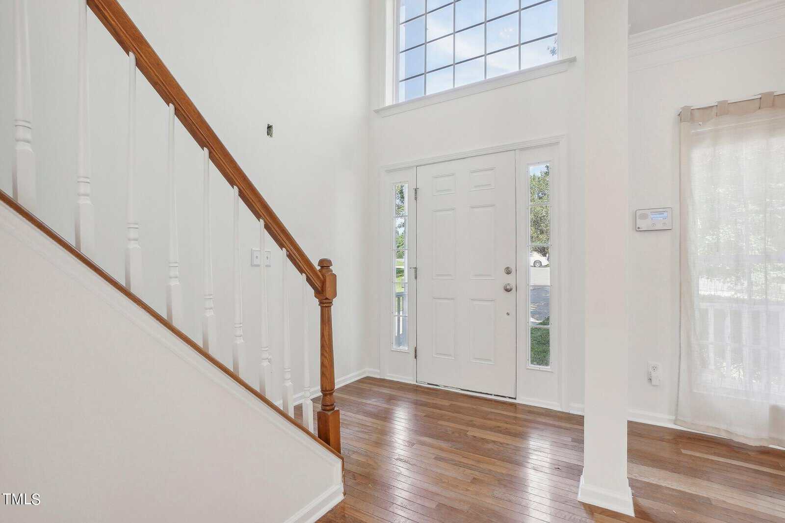 Bright entryway featuring a wooden staircase with white balusters. The space has a wooden floor and high ceiling, with a large window and a white front door flanked by narrow glass panels. Natural light streams in, creating a welcoming atmosphere.