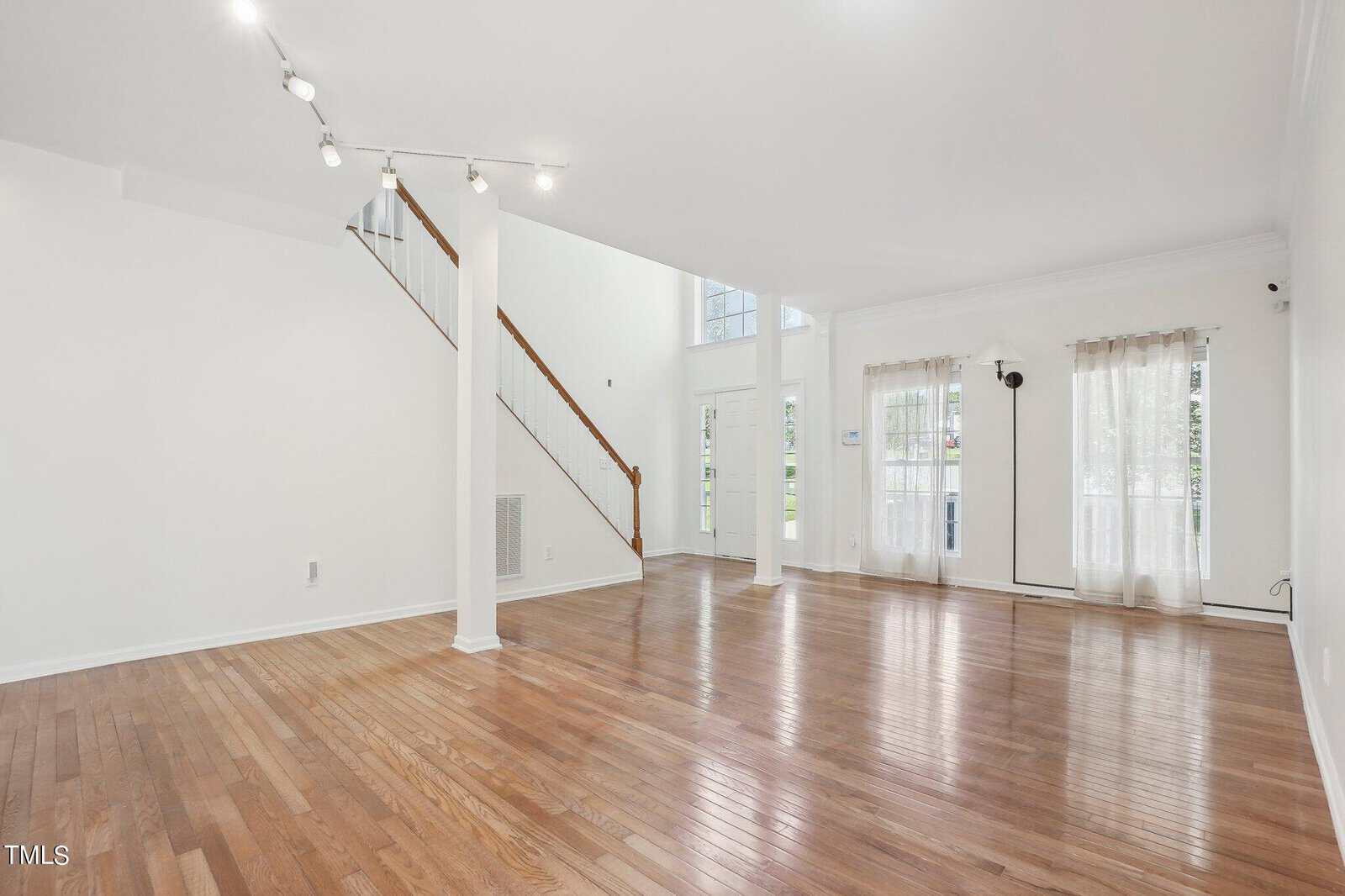 Spacious empty living room with polished hardwood floors and tall white walls. Large windows with sheer curtains allow natural light to fill the space. A staircase with wooden railing leads to the upper level. Ceiling spotlights illuminate the area.