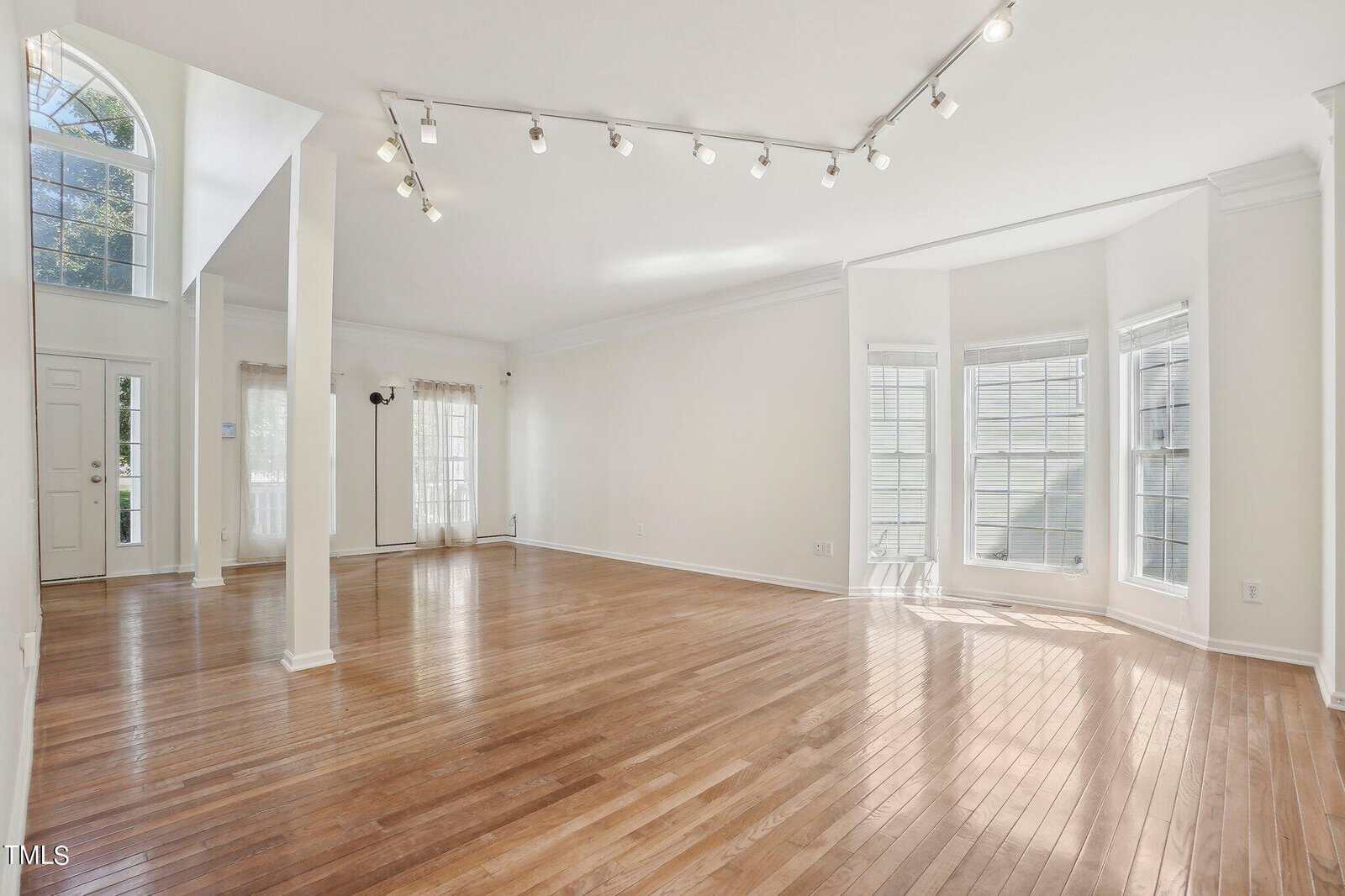 Spacious living room with hardwood floors, large windows, and white walls. Ceiling track lighting and a high, arched entrance with additional windows bring in natural light. Open floor plan leading to other areas.