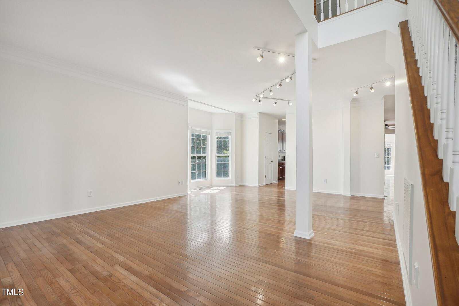 A spacious, empty living room with polished hardwood floors and white walls. Sunlight streams in through large windows. A staircase with a wooden handrail is on the right, and track lighting is on the ceiling.