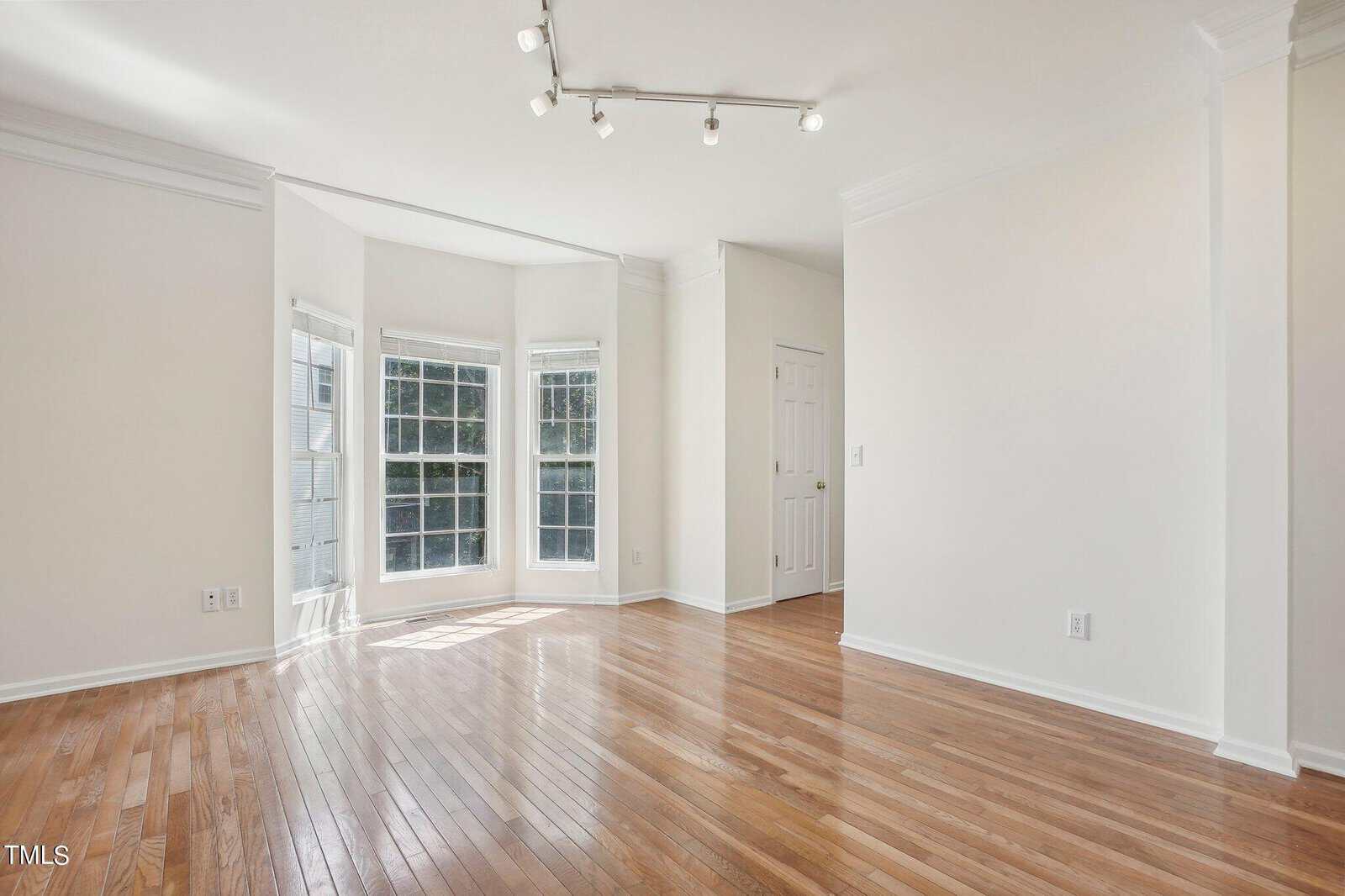A bright, empty room with large windows and wooden floors. The walls are white, and there is a door in the corner. Ceiling lights are positioned above, creating a clean, open space with abundant natural light.