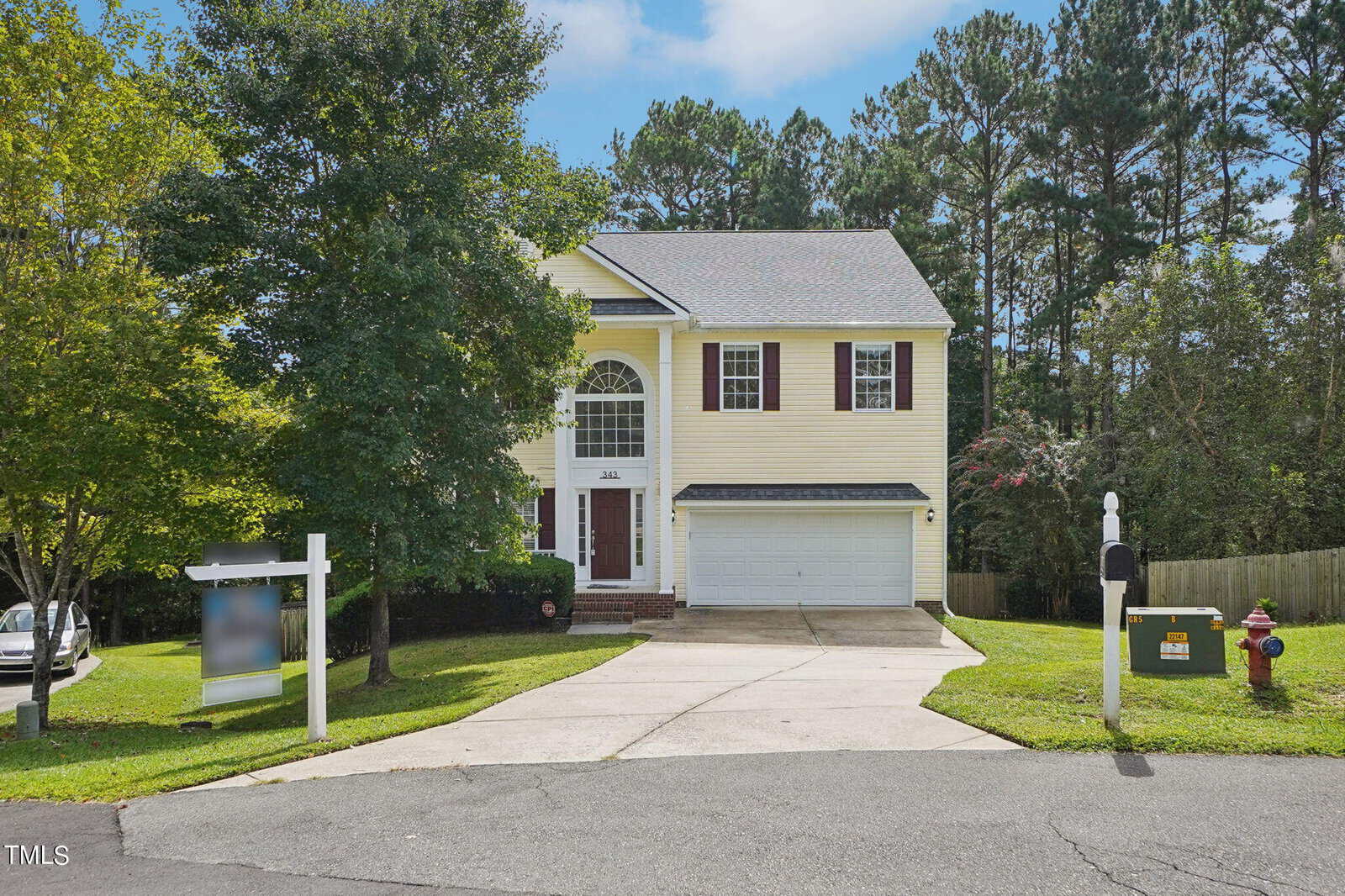 A yellow two-story house with a black garage door and white trim, located on a cul-de-sac. It has a concrete driveway, a mailbox near the road, and trees surrounding the property. A "For Sale" sign is visible on the left of the driveway.