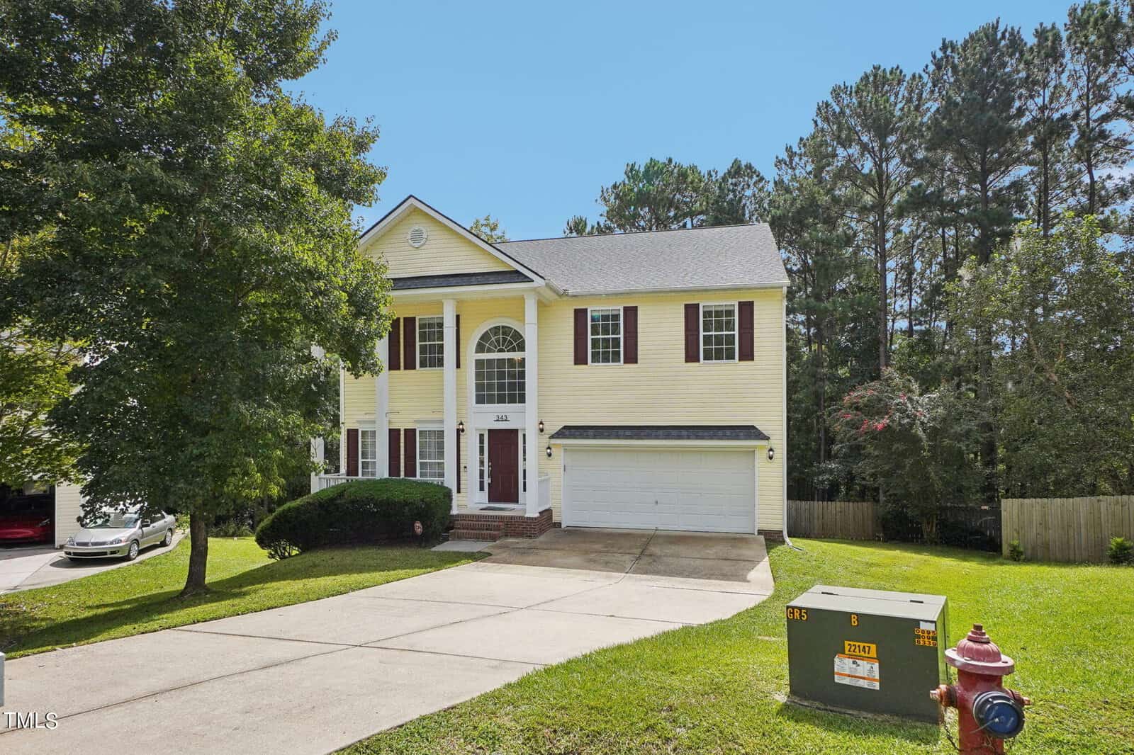 A charming two-story yellow house at 343 Howland Court, with brown shutters and a brick front porch, sits gracefully on a well-maintained lawn with trees in Creedmoor. A driveway leads to a double garage, while a fire hydrant and utility box stand in the foreground.