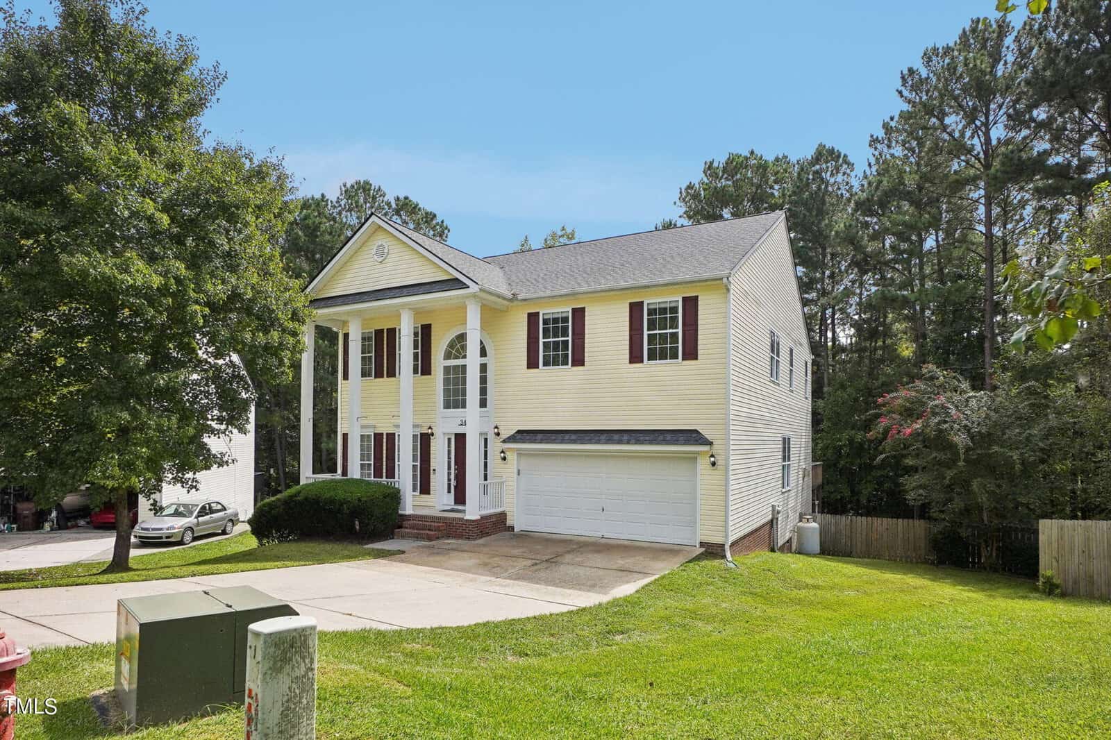 A two-story yellow house with white columns and red shutters is situated in a grassy yard. It features a double garage, a gabled roof, and a driveway. The background displays a row of trees and a bright blue sky.