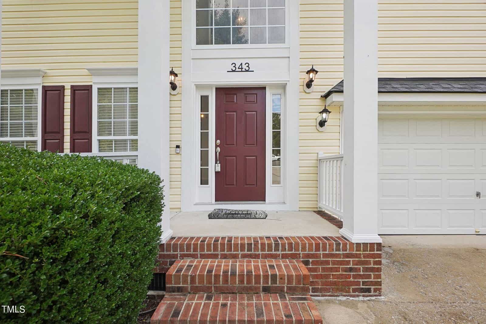 A red front door with a transom window is centered between two white columns and brick steps. Flanked by lights, the door is numbered "343." Adjacent are large windows with shutters and a white garage door on a yellow house. A green shrub adds contrast.